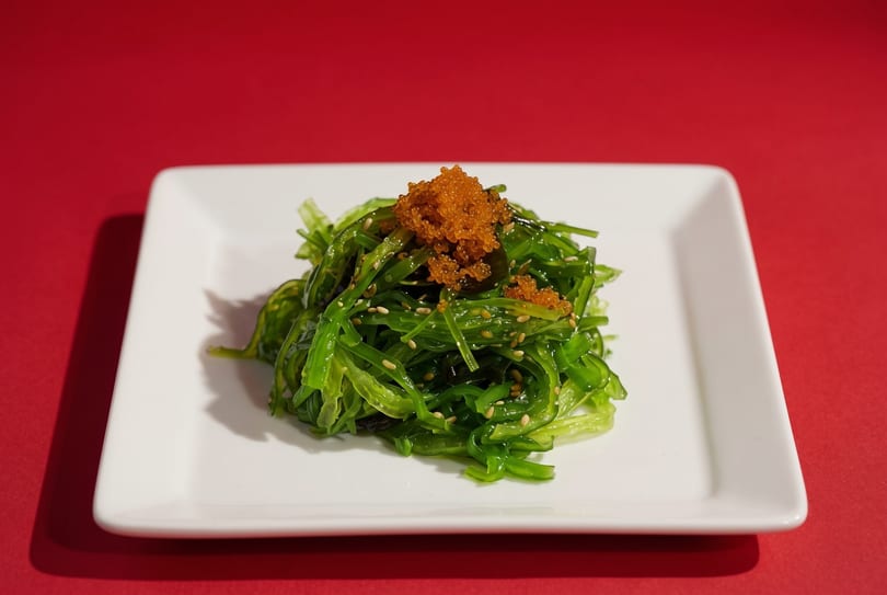 Bright green seaweed salad with orange tobiko and sesame seeds on a white rectangular plate.