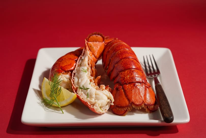 Two cooked lobster tails, one split open, with a lemon wedge and dill on a white plate.