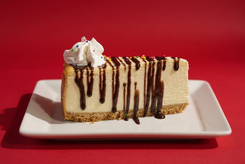 Cheesecake slice with chocolate drizzle, whipped cream, and sprinkles on a white plate.