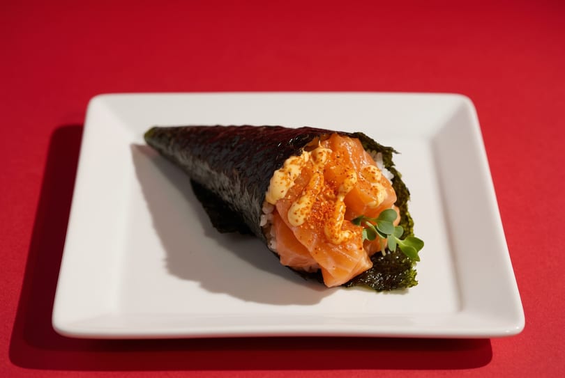 Salmon temaki hand roll with spicy mayo and microgreens on a white plate against a red background.