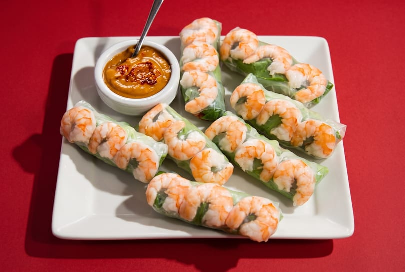 Shrimp spring rolls with peanut dipping sauce on a white rectangular plate against a red background.