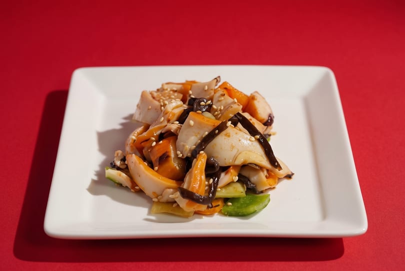 A white rectangular plate holds a colorful squid salad with vegetables and sesame seeds on a red background.