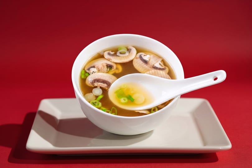 Mushroom soup with green onions in a white bowl and spoon on a white plate against a red background.