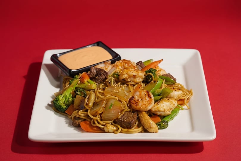 A white rectangular plate holds a stir-fry of noodles, shrimp, beef, and vegetables with a side of sauce.