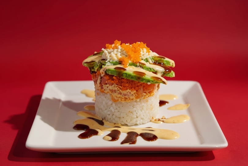 Sushi stack with rice, tuna, avocado, crab, and roe, drizzled with sauces on a white plate.