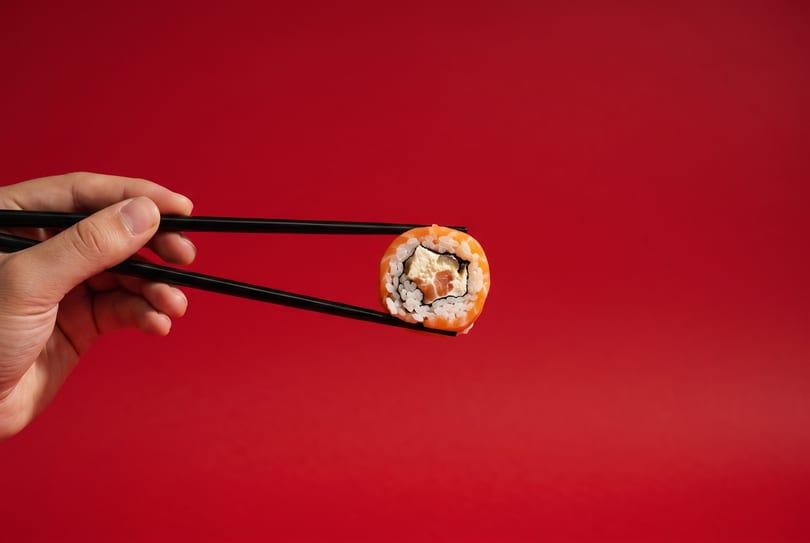 A hand holds a single sushi roll with salmon and cream cheese using black chopsticks against a red background.