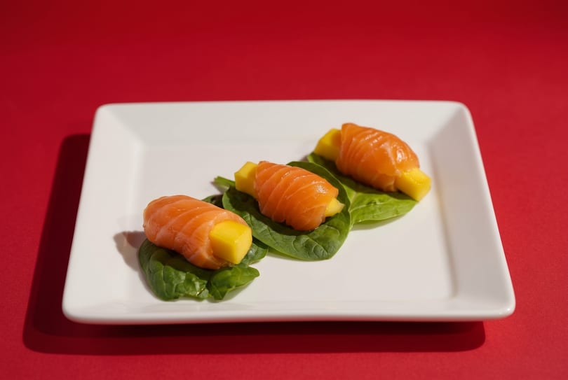 Three salmon and mango rolls on spinach leaves, arranged on a white rectangular plate.