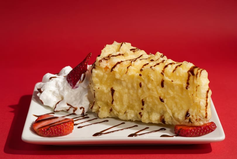 Tempura cheesecake with chocolate drizzle, whipped cream, and fresh strawberries on a white plate.
