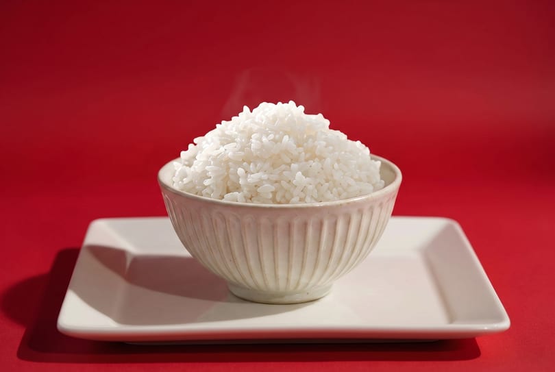 Steaming white rice piled high in a textured bowl, centered on a white rectangular plate against a red background.