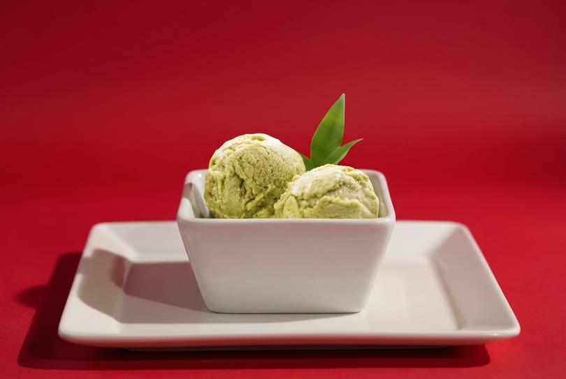 Two scoops of green tea ice cream with a green leaf garnish in a white bowl on a red background.