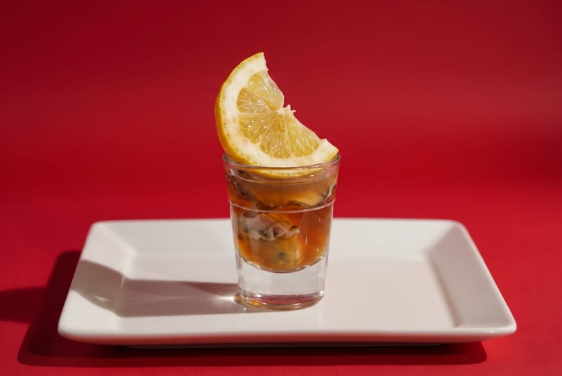Oysters in a shot glass with a lemon slice, on a white rectangular plate against a red background.