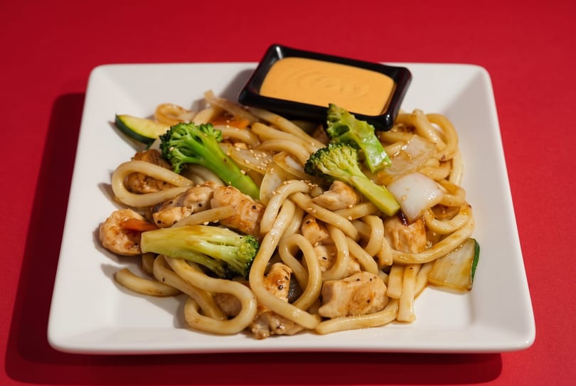 Udon noodles with chicken, shrimp, broccoli, zucchini, and onions, served with a side of sauce.